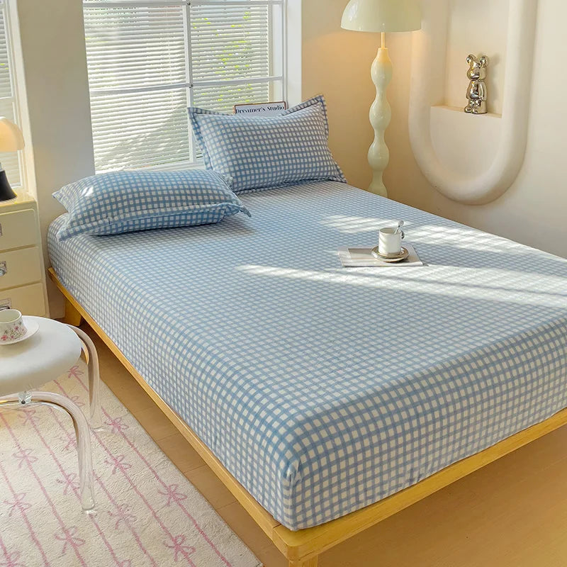 A light blue plaid flannel fitted sheet is laid on a bed with white bedding and a pink patterned rug.