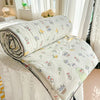 A white quilt blanket with a pastoral print featuring yellow and green floral designs is rolled up on a white surface.