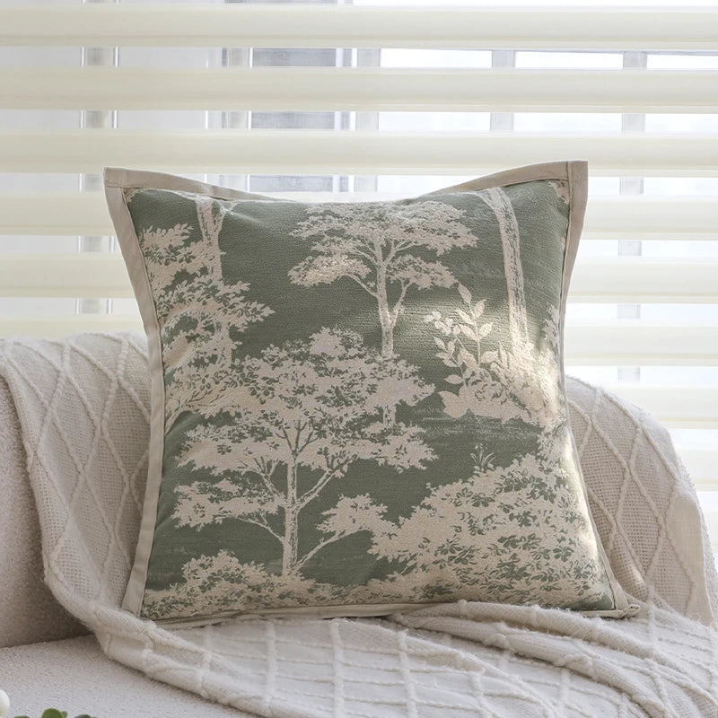 A square cushion cover with an embroidered floral pattern in sage green and cream tones, resting on a light-colored textured 