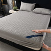 A gray quilted mattress protector is shown on a bed, with a hand touching the fabric.