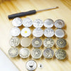 A collection of 30 silver jeans buttons laid out on a wooden surface with a screwdriver nearby.