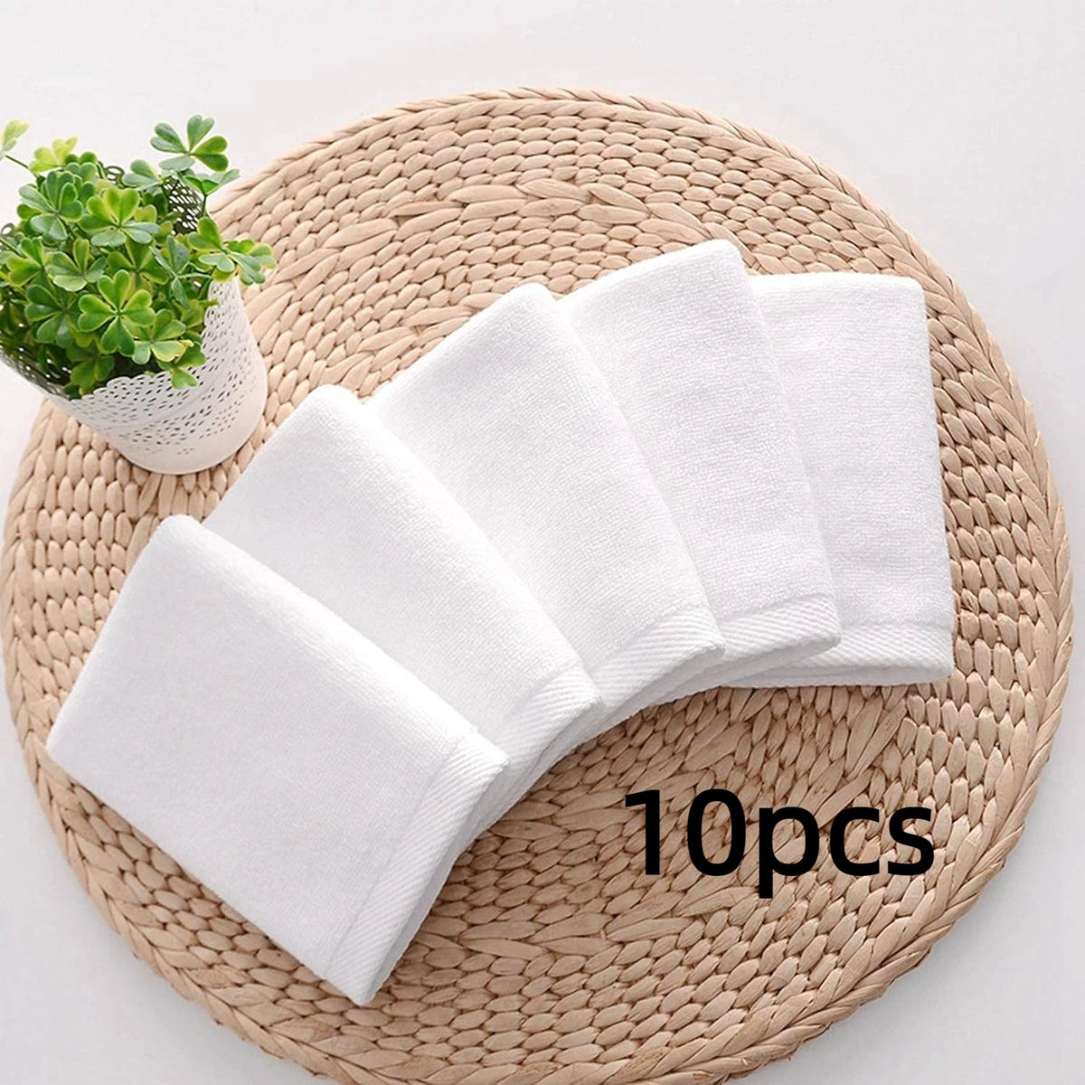 A set of ten white cotton face towels is displayed on a woven mat next to a small potted plant.