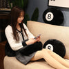 A young woman is sitting on a sofa holding a smartphone, next to a black 8 ball plush pillow.