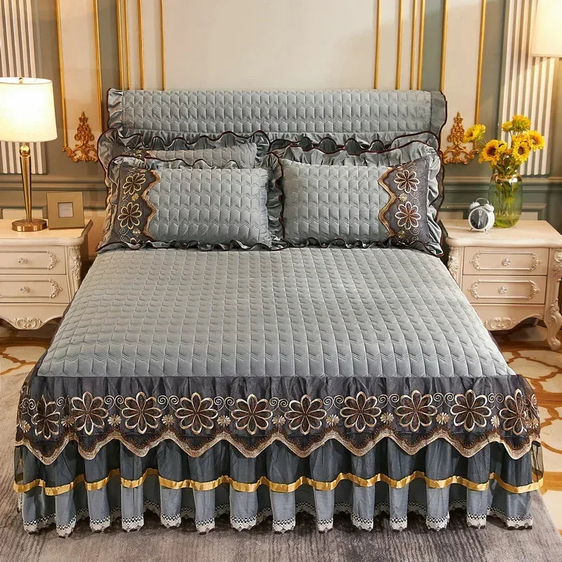 Gray quilted bed skirt with dark floral embroidery and gold trim on a bed.