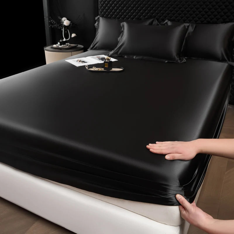 A hand is adjusting a black satin fitted sheet over a mattress.