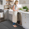 A woman stands on a gray anti-fatigue PVC kitchen mat in a bright kitchen with white cabinets and countertops.