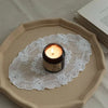 A small candle sits on a white lace coaster placed on a beige ceramic plate.