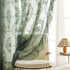 A sheer floral lace curtain panel in sage green hangs beside a light-colored upholstered chair.