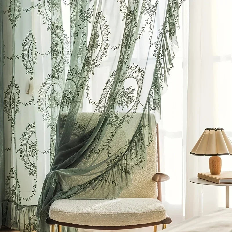 A sheer floral lace curtain panel in sage green hangs beside a light-colored upholstered chair.