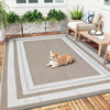 A light brown dog lies on a gray outdoor patio rug next to wooden decking and potted greenery.