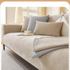 A beige waterproof anti-scratch sofa protection cover is shown on a bed with light gray pillows and natural light.
