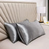 A set of smooth, silver satin silk pillows resting against a light upholstered headboard with a bedside lamp.