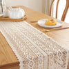 A white crochet lace table runner with fringe is displayed on a wooden table next to a teapot and a small pastry.