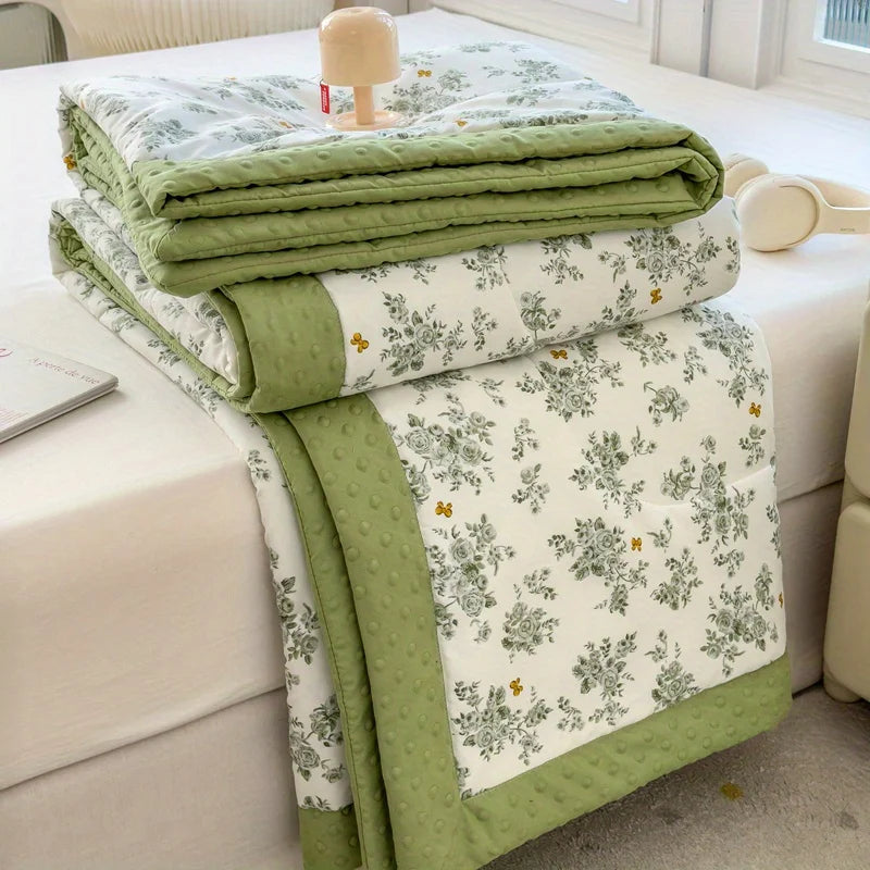 A stack of light green floral printed summer quilts folded on a white bed sheet.