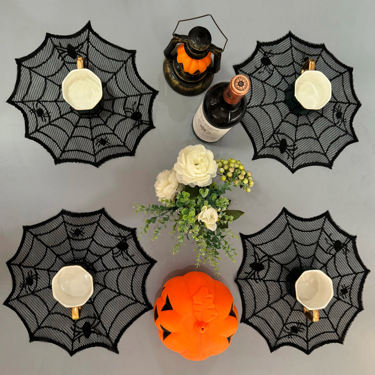 A black spider web design features several white ceramic cups and an orange pumpkin on a gray surface.