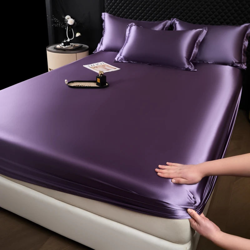 A purple satin fitted sheet is draped over a mattress, with a hand adjusting the fabric.