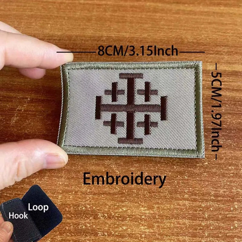 A close-up of a square embroidered patch featuring a cross design on a green fabric, shown next to a hand and a sewing tool.