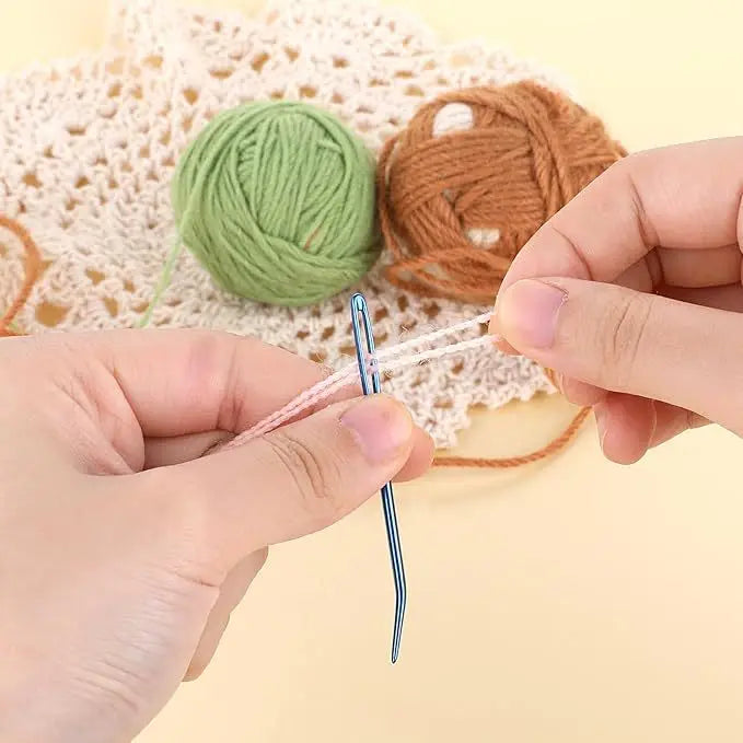 A pair of bent tip tapestry needles is shown being used to thread a green yarn onto a piece of twine.