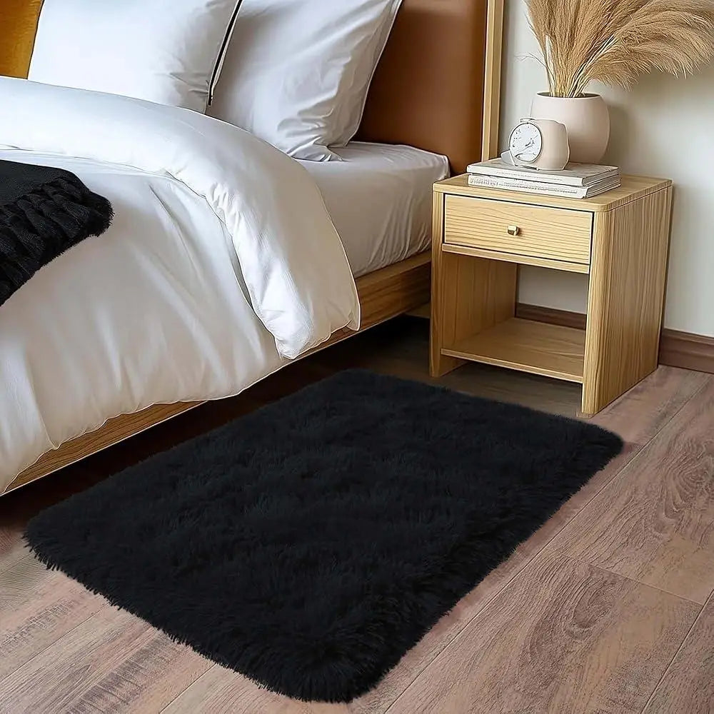 A plush solid black area rug placed on a wooden floor next to a bed and bedside table.