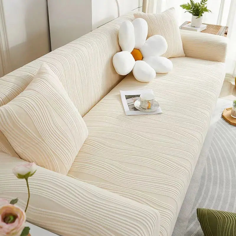 A beige stretchable anti-slip sofa cover with white decorative pillows and a small tray on a light-colored sofa.