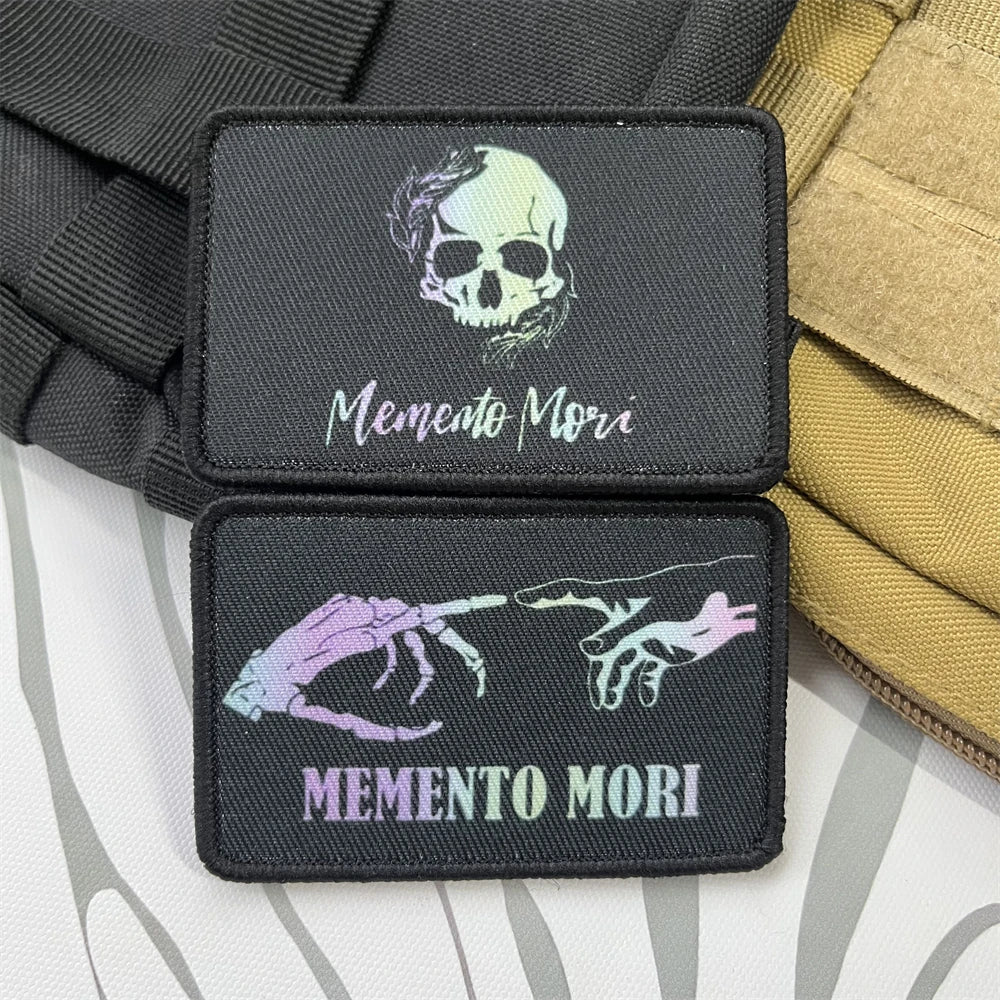 Two black badge patches featuring a skull design and the text Memento Mori on a tactical strap.
