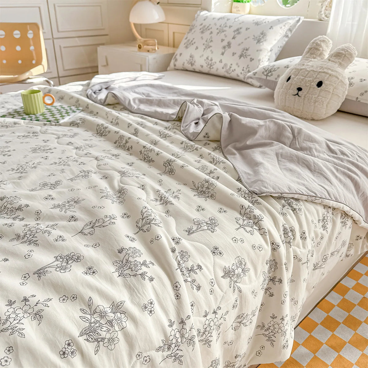 A white quilt blanket with a delicate floral print is displayed on a bed, next to a cream-colored stuffed animal.