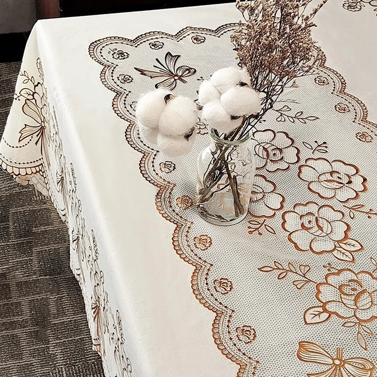 A white dining table cloth with a lace pattern is set with dried floral arrangements and fluffy white pom-poms.