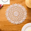 A white floral embroidered round placemat is displayed on a wooden table next to a plate.