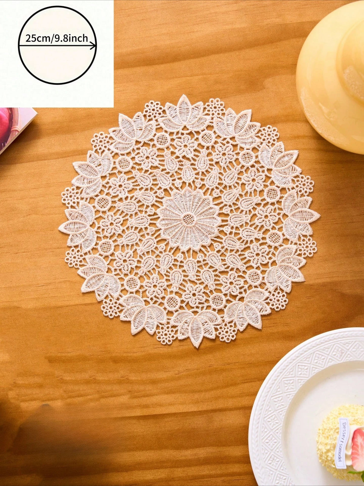 A white floral embroidered round placemat is displayed on a wooden table next to a plate.