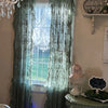 A pair of sheer floral lace curtains in a muted sage green color hanging over a window.