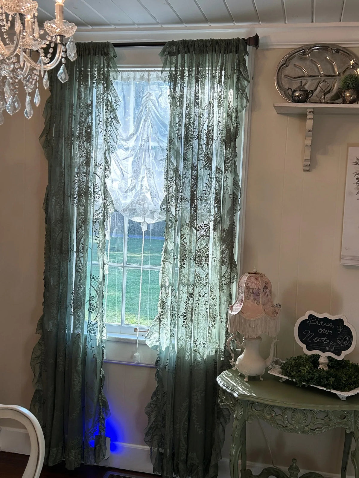A pair of sheer floral lace curtains in a muted sage green color hanging over a window.