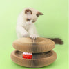 A small white kitten sits next to a round, textured cardboard cat scratching board set.