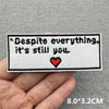A black embroidered patch with the text Despite everything it's still you and a red heart on a gray fabric background.