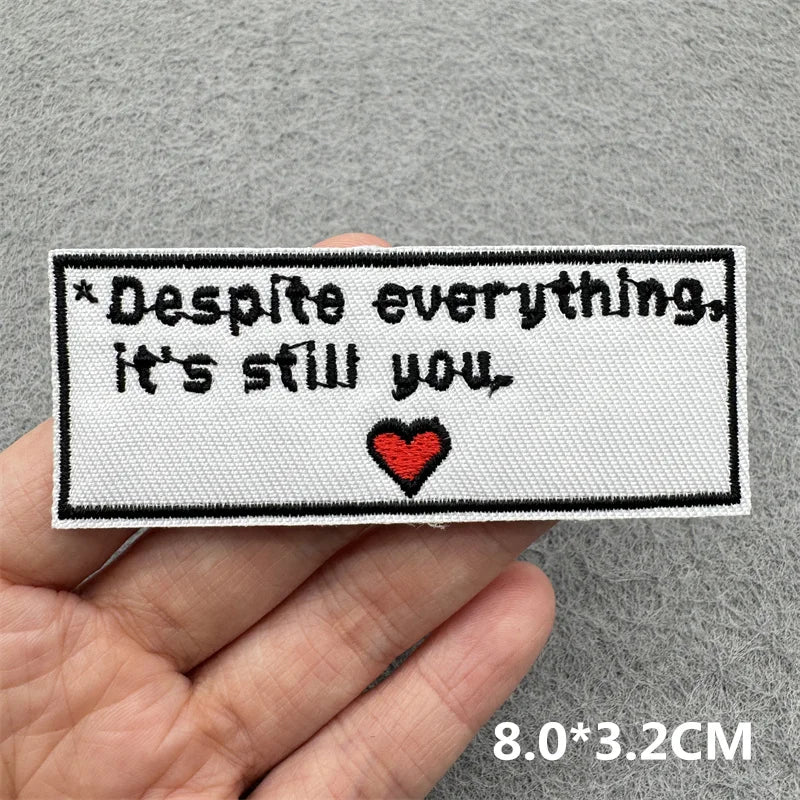 A black embroidered patch with the text Despite everything it's still you and a red heart on a gray fabric background.