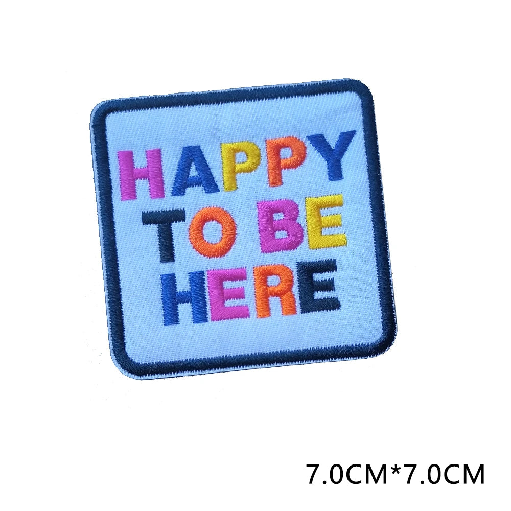 A square embroidered patch with the text HAPPY TO BE HERE in colorful thread on a blue fabric background.
