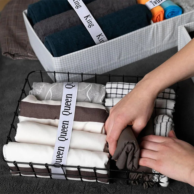 A hand is organizing rolled white and gray bed sheets in a black wire basket.