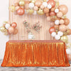 An orange sequin rectangle table cover is draped on a wooden surface, set against a backdrop of pink and gold balloons.