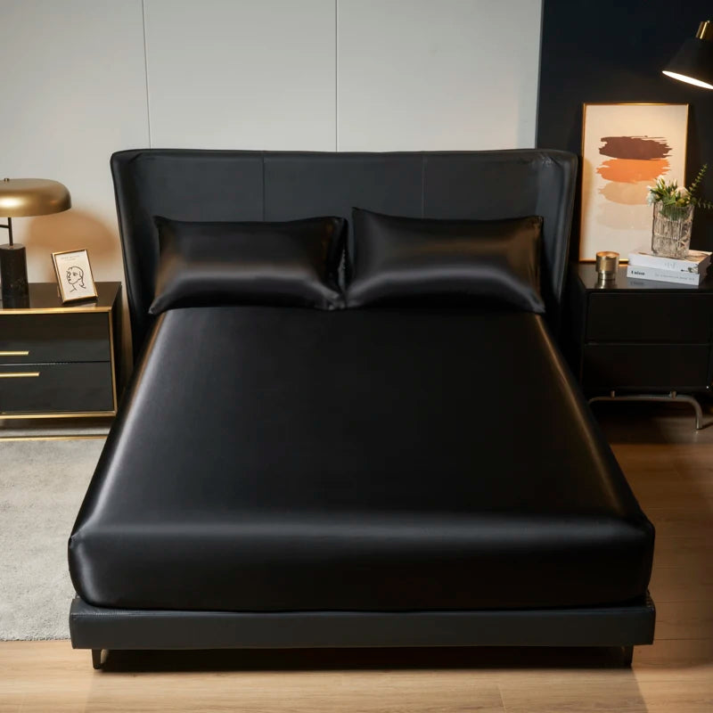 A black satin bedding set with an elastic band is displayed on a modern bed in a well-lit room.