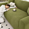 A person is relaxing on a moss green stretch jacquard sofa slipcover with a white knitted pillow and a cup of tea.