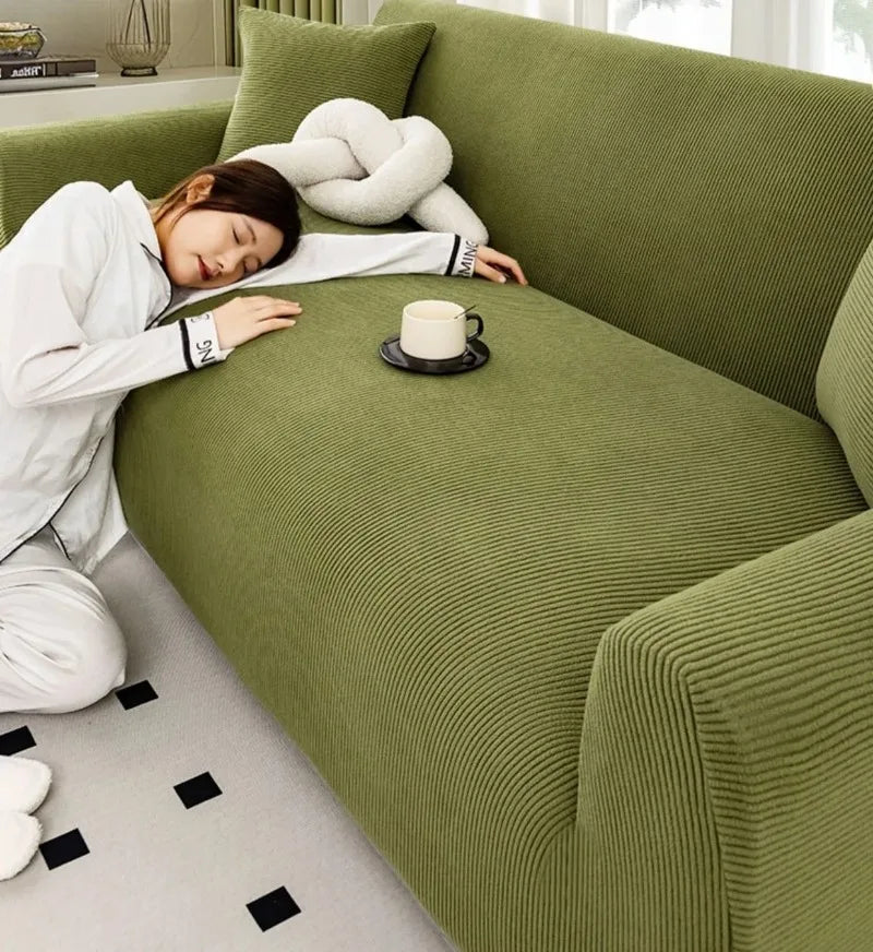 A person is relaxing on a moss green stretch jacquard sofa slipcover with a white knitted pillow and a cup of tea.