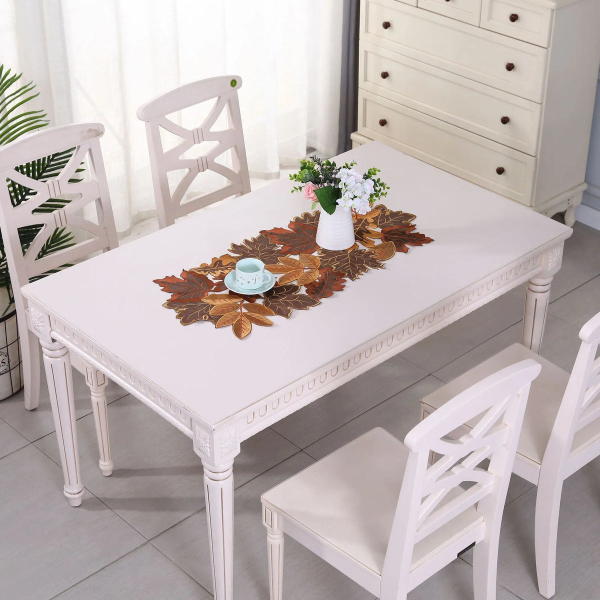 A white farmhouse table is set with a maple leaf runner and floral centerpiece, surrounded by white wooden chairs.
