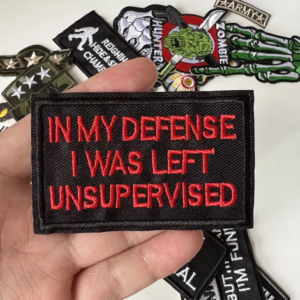 A hand holds a black embroidered patch with red text that reads IN MY DEFENSE I WAS LEFT UNSUPERVISED.