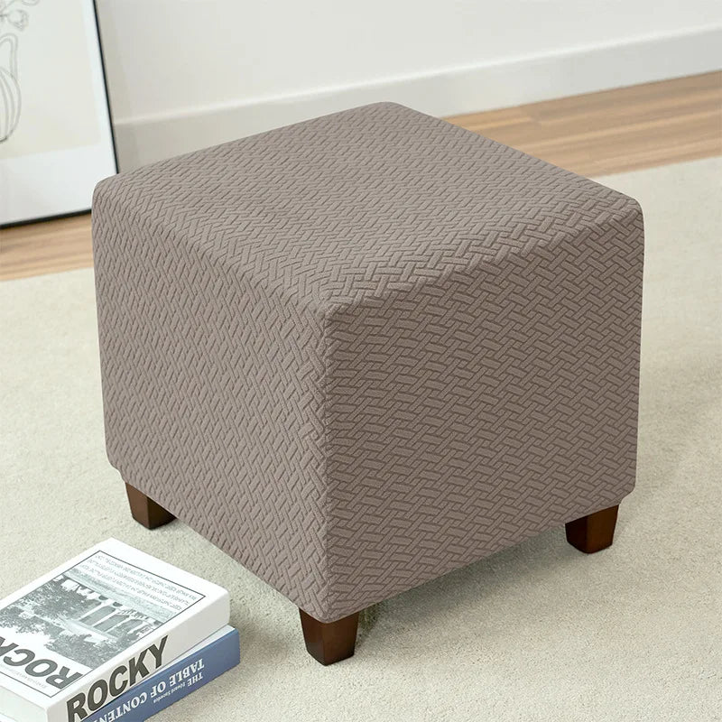 A square taupe jacquard stretch ottoman slipcover resting on a light beige carpet.