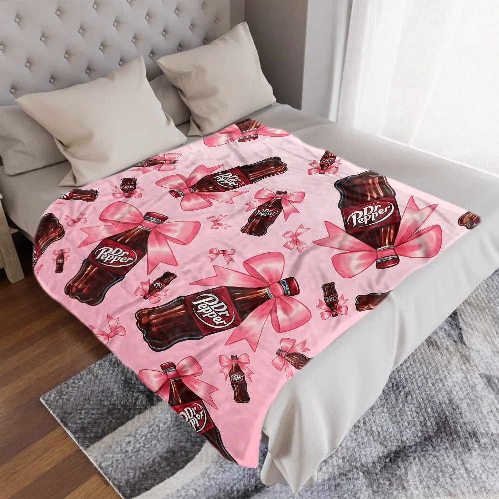 A pink Dr. Pepper print cozy blanket is displayed on a bed with white linens and a gray throw.
