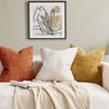 A close-up of a beige textured throw pillow cover resting on a light-colored sofa with a framed botanical print in the backgr