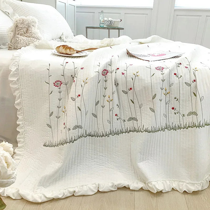 White quilt bedspread with delicate floral embroidery featuring pink and green stems on a light wood surface.