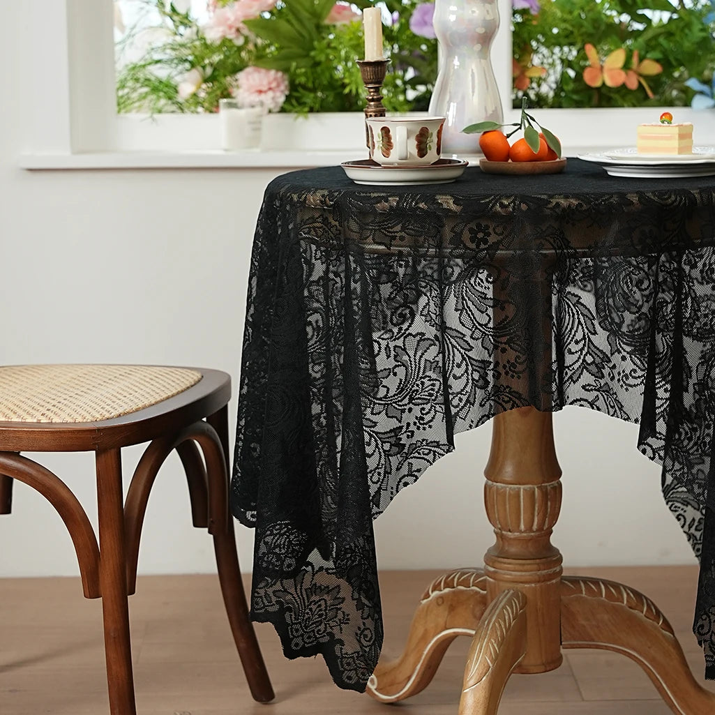 A black lace tablecloth is draped over a wooden table, set for dining with fruit and a candle.