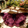 A deep purple irregular shaped plush floor rug is displayed on a wooden table in a bright, sunlit indoor garden setting.