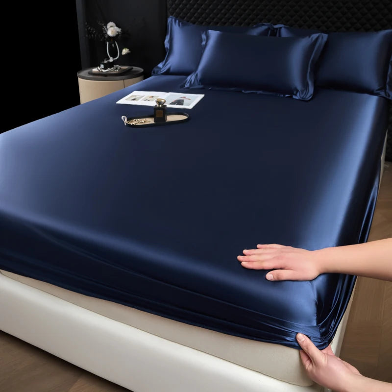 A dark blue satin fitted sheet is draped over a mattress, with a hand adjusting the fabric.