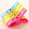 A pack of four large plastic clothes pins in blue, yellow, and pink are shown on a wooden surface.